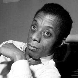 James Baldwin, holding a cigarette, sits in front of a typewriter looking intently ahead.