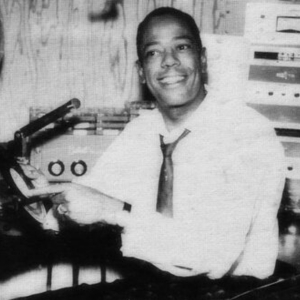 Jocko Henderson smiles as he holds a record, seated in front of his radio broadcast desk.