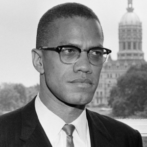Malcolm X, in a suit and his trademark glasses, is photographed looking stern and standing in front of a window.