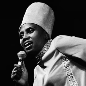 Miriam Makeba sings into a microphone wearing a sparkling outfit and tall hat.