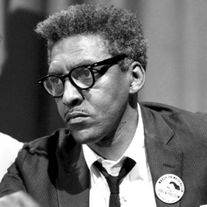 A photo of Bayard Rustin shows a man with graying hair, a mustache, black glasses, and a suit, looking gravely ahead.