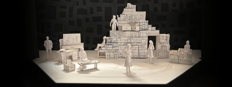 A white, cardboard scenic model for STOKELY: THE UNFINISHED REVOLUTION. A bunch of shelves and drawers make up a large tower in the center of the stage, and there are smaller structures on either side.