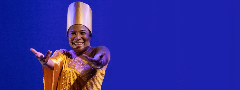 A person in a gold dress and tall, gold hat smiles and stretches their hands to the audience.