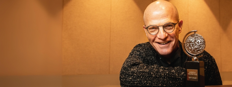 A man smiles in front of a Tony Award.