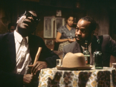 The interior of a juke joint. Two men are sitting at a table - one is speaking, one is listening intently - and there is a woman in the background.