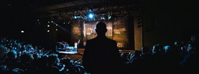 A photo of a man watching a performance onstage. The photo is taken from behind, so the only thing visible is the man's silhouette.