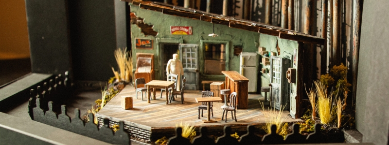 A set model of the interior of a juke joint. There are two tables and some chairs and stools, a bar in the corner, and colorful posters on the walls. The building looks run-down.