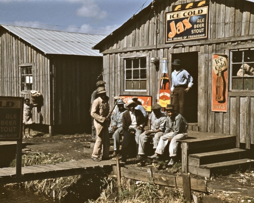 A simple wooden building with signs on the exterior walls of a juke joint advertising beer and Coca-Cola. Several Black men sit on the open porch, wearing hats.