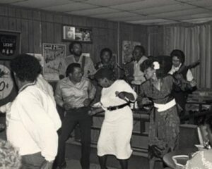 A black and white image of a band playing onstage while 4-5 people dance exuberantly below the stage.