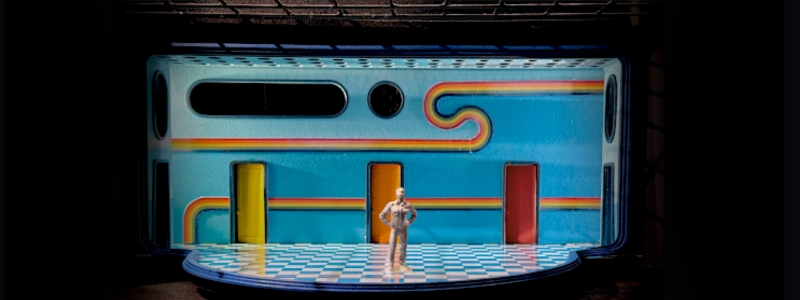 A scenic model for the set of FALSETTOS. There is a bright blue background with a rainbow ribbon wending its way around the set, there are three colorful doors, a checkerboard floor, and a figure standing in the center.