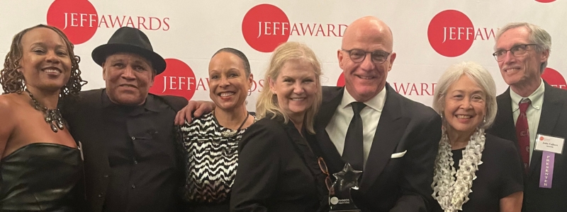 A group of people stand in front of a background with text for the Jeff Awards. Someone is holding a clear award.