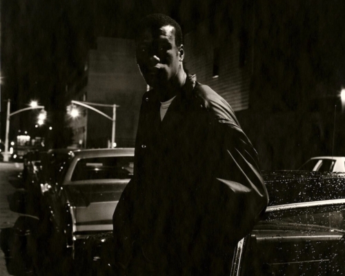 A man in a dark jacket leans against a car dotted with raindrops.