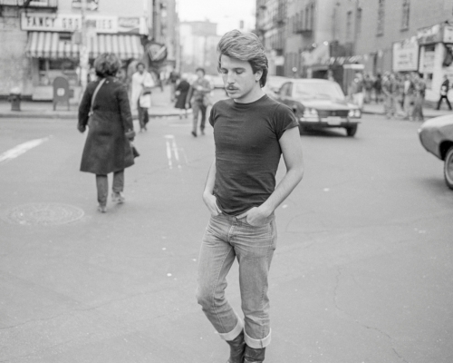 A man with windswept hair walks down the street with his hands in his pockets in 1976 New York City.