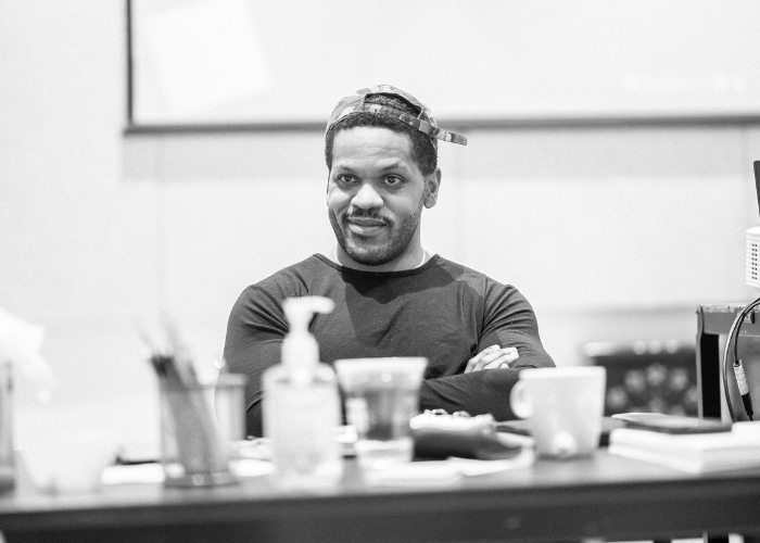 A person sits behind a table. They are smiling with their arms crossed in front of them.