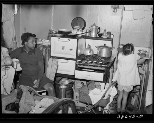 Two children in a crowded kitchennette, surrounded by piles of household supplies and miscellaneous items. The kitchenette is cramped and busy.