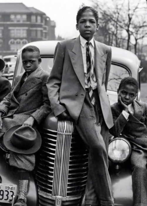 Five Black boys wearing suit jackets and ties sit on top of or lean against the hood of a fancy car. A Chicago boulevard is visible behind them.