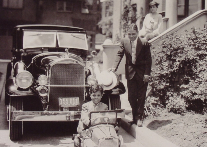 A young Black boy sits smiling in a toy car that replicates the vehicle behind him. His father and mother watch from behind him. The family appears to be quite wealthy - they are outside of a nice home and wearing expensive clothes.