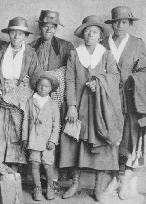 A family of eight Black folks, including a young child and some elders, poses for a photo after recently migrating to Chicago from the American South. They carry and wear traveling cloaks. Two suitcases sit in the foreground.