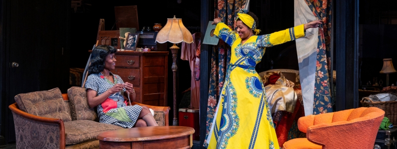 A woman presents herself from behind a curtain, joyously and with arms spread. She is wearing a bright yellow and blue dress and head wrap, and showing it off to another woman who is sitting on the couch, knitting.