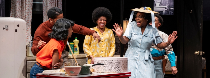 A group of people smiling and dancing. One of the people is striking a pose and wearing a gardening hat with blue ribbons.